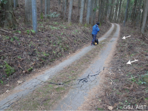 写真1-1：持山の南東方の舗装道路を斜めに横切る開口断裂群（矢印）。北西方向を見る。（地点1）